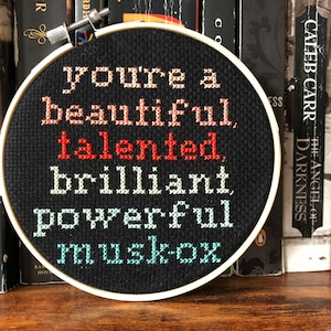May include: A black cross-stitch embroidery hoop with the words "you're a beautiful, talented, brilliant, powerful musk-ox" in various colors. The hoop is displayed in front of a bookshelf filled with books.