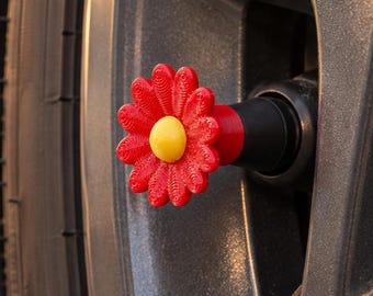 May include: A red and yellow flower-shaped valve stem cap attached to a dark gray car wheel rim. The decorative cap features a textured red petal design surrounding a smooth yellow center, securely fastened to the black tire valve stem.