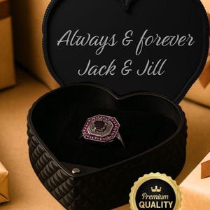 May include: A black heart-shaped ring box with the words "Always & forever Jack & Jill" in white script. Inside, a silver ring with a square, ruby-colored center stone and pink accents. A gold "Premium Quality" sticker is visible.