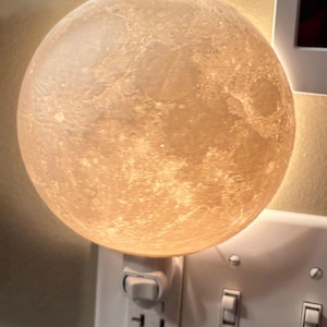 May include: A glowing, spherical night light shaped like the moon, plugged into a white wall outlet. The moon has a textured surface with craters and is lit from within, casting a warm, soft light. The outlet and light switches are also visible.