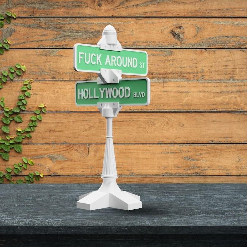 May include: A white street sign with two green signs that read "FUCK AROUND ST" and "HOLLYWOOD BLVD". The sign is on a white post with a decorative base. The background is a wooden wall.