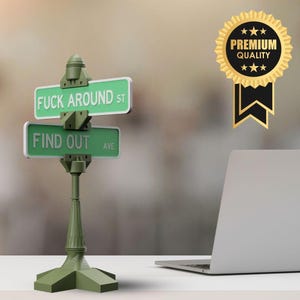 May include: A green street sign with the phrases "FUCK AROUND ST" and "FIND OUT AVE" in white text. The sign is mounted on a green post. A gold and black "PREMIUM QUALITY" badge is also visible.