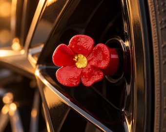 May include: A bright red flower-shaped valve stem cap with a textured yellow center, attached to the black metal rim of a vehicle wheel. The five-petaled floral design is made of a solid material and is positioned centrally on the wheel hub.