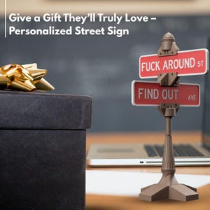 May include: A personalized street sign with the text "FUCK AROUND ST" and "FIND OUT AVE" in white on a red background. The sign is mounted on a brown decorative post. A black gift box with a gold bow is in the background.