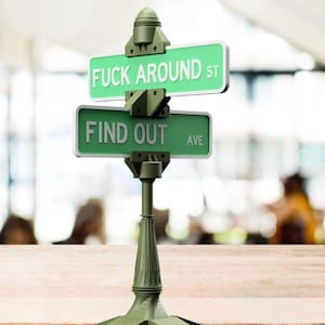 May include: A green street sign with the text "FUCK AROUND ST" above "FIND OUT AVE". The sign is mounted on a decorative green post with a base. The background is blurred.