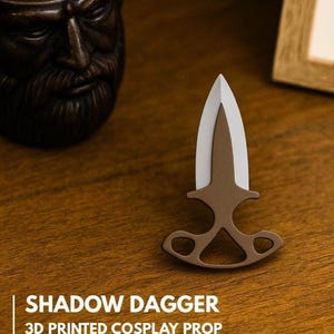 Puede incluir: Una daga de sombra impresa en 3D, accesorio de cosplay marrón y blanco, con un mango decorativo. El texto "SHADOW DAGGER" es visible. La daga se muestra sobre una superficie de madera.