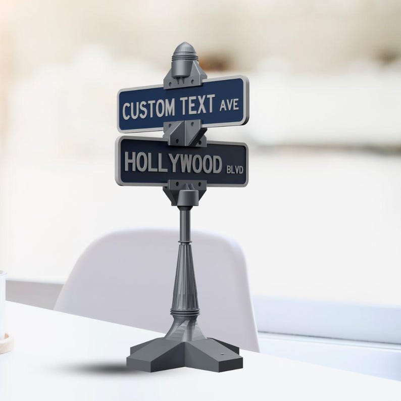May include: A gray street sign with two blue rectangular plaques. The top plaque reads "CUSTOM TEXT AVE" and the bottom plaque reads "HOLLYWOOD BLVD". The sign is mounted on a gray post with a decorative base.