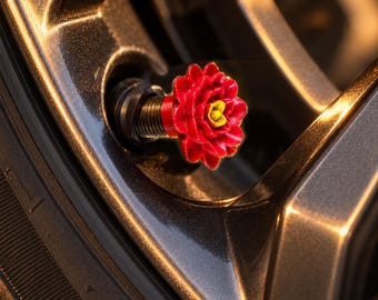 May include: A red and yellow flower-shaped tire valve cap attached to a dark metallic car wheel rim. The valve cap features a layered petal design in bright red with a small yellow center, mounted on a threaded black metal stem base.