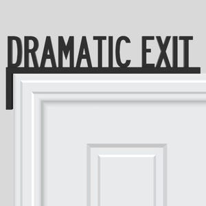 May include: Black metal sign that reads "DRAMATIC EXIT" mounted above a white door. The sign is in a bold, sans-serif font and is designed to be mounted on the top of a door frame. The door has a recessed panel design.