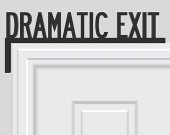 May include: Black metal sign that reads "DRAMATIC EXIT" mounted above a white door. The sign is in a bold, sans-serif font and is designed to be mounted on the top of a door frame. The door has a recessed panel design.