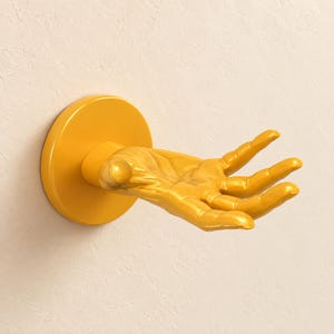 May include: A bright yellow wall-mounted hook shaped like an open human hand. The design features a circular base plate attached to a sculpted hand with fingers extended upward, providing a functional surface for hanging items on a wall.