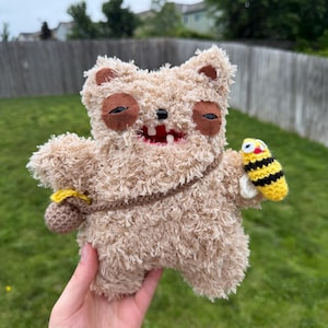 May include: A fuzzy beige crochet creature with dark brown felt eye patches, a wide mouth showing white teeth, and a brown strap across its chest. It carries a small brown pouch on one side and holds a tiny yellow and black striped bee toy.