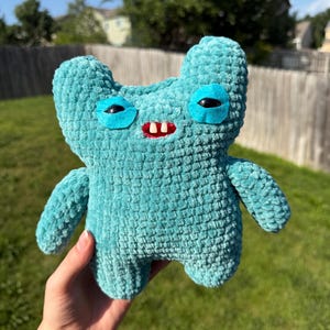 May include: A turquoise plush toy made of soft, textured yarn. The creature features two rounded ears, two black plastic eyes set within bright turquoise felt patches, and a small red mouth with two prominent white teeth protruding from the center.
