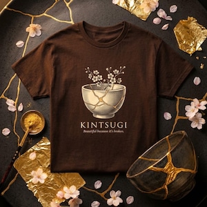 Può includere: Una maglietta marrone con un design Kintsugi, raffigurante una ciotola rotta con fiori di ciliegio. Il testo recita "Kintsugi Beautiful because it's broken". Foglie d'oro e fiori di ciliegio circondano la maglietta.