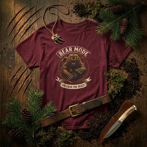Può includere: Una maglietta bordeaux con una grafica di orso e la scritta "BEAR MODE UNLEASH THE BEAST". L'immagine include anche una cintura in pelle, un coltello e una collana con un ciondolo a forma di artiglio. Gli oggetti sono disposti su una superficie di legno.