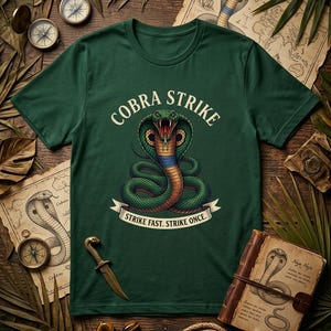 Può includere: T-shirt verde scuro con grafica di un cobra. La scritta "COBRA STRIKE" è sopra il cobra, e "STRIKE FAST. STRIKE ONCE." è sotto. Il cobra è verde, rosso e blu. La maglietta è su una superficie di legno con mappe e bussole.