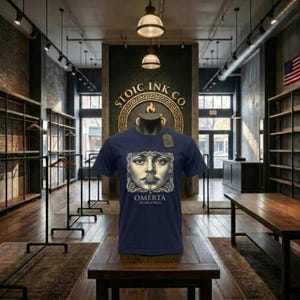 Può includere: T-shirt blu navy con una grafica dettagliata di un volto e la parola "OMERTA" in bianco. Il testo "The Code of Silence" è sotto. La maglietta è esposta su un manichino in un negozio.