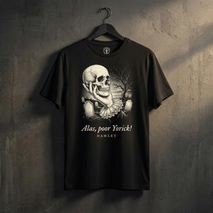Könnte beinhalten: Ein schwarzes Kurzarm-T-Shirt mit einem Grafikdruck einer Hand, die einen menschlichen Schädel auf einem Friedhof hält. Der Text "Alas, poor Yorick!" und "HAMLET" ist in Weiß unter der Illustration gedruckt. Das T-Shirt hängt an einem Kleiderbügel.