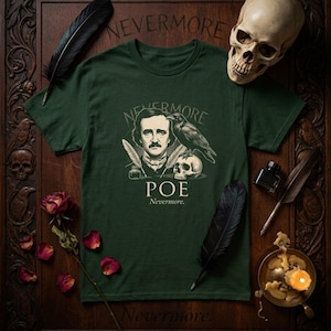 Può includere: T-shirt verde scuro con un ritratto di Edgar Allan Poe, un corvo e un teschio, con le parole "Nevermore" e "POE". La maglietta è circondata da piume, un teschio e petali di rosa.