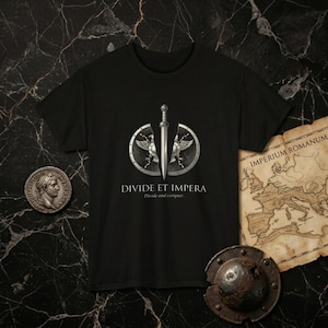 May include: A black short-sleeved t-shirt featuring a graphic of a silver sword positioned vertically between two halves of a shield with eagle wings. Below the graphic, the text reads DIVIDE ET IMPERA and Divide and conquer.