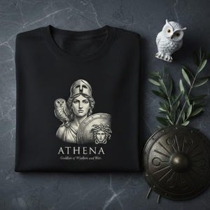 May include: Black crew neck sweatshirt featuring a detailed illustration of Athena, the Greek goddess, with an owl and shield. The text "ATHENA Goddess of Wisdom and War" is printed below the image. A small white owl figurine and a decorative shield are also visible.