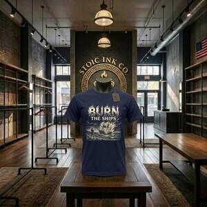 Può includere: T-shirt blu navy con la scritta "BURN THE SHIPS" sopra un'illustrazione dettagliata di una nave in mare. La maglietta è esposta su un manichino in un negozio, con il logo "STOIC INK CO." visibile sullo sfondo.