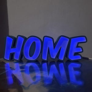 Op de afbeelding: Een blauw LED-lichtbord met het woord "HOME". De letters zijn omlijnd in een donkerdere blauwe kleur, met een heldere blauwe gloed die van binnenuit komt. Het bord staat op een reflecterend oppervlak.