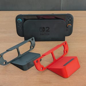 Könnte beinhalten: Eine schwarze Nintendo Switch Konsole und Dockingstation mit zwei Controller-Haltern, einer grauen und einer roten. Das Dock hat das Nintendo Switch Logo. Die Halter sind zur Aufnahme der Controller konzipiert.