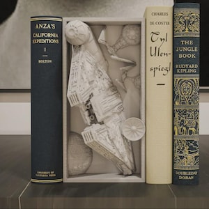 Puede incluir: Una exhibición de libros con un modelo 3D de una nave espacial dentro de una caja. Los libros incluyen "Anza's California Expeditions", "Tyl Uilenspiegel" y "The Jungle Book". Los libros son de varios colores, incluyendo azul marino y crema.
