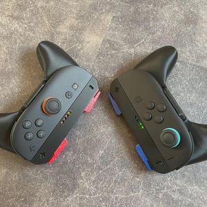 Puede incluir: Dos mandos de juego negros con empuñaduras negras adjuntas. Uno tiene detalles en rojo, el otro en azul. Cada mando tiene un joystick, botones y una cruceta. Los mandos están sobre una superficie gris.