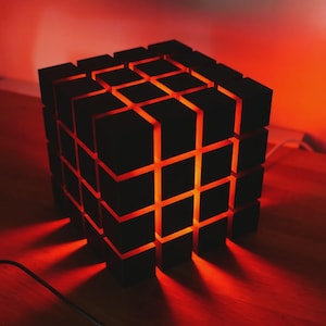 Può includere: Lampada a forma di cubo nero con un motivo a griglia. L'interno emette una calda luce arancione, in contrasto con l'esterno scuro. Il design della lampada proietta ombre sulla superficie in legno, accentuando la forma geometrica.