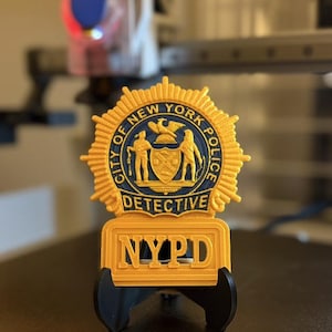 以下が含まれることがあります： 金色のNYPD探偵バッジ。青と金のエンブレムが付いています。バッジには「City of New York Police」と「Detective」の文字が記されています。「NYPD」の文字がバッジの下部に表示されています。