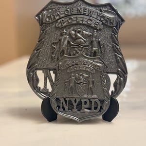 以下が含まれることがあります： 銀色のNYPD警察バッジ。盾型で、複雑なディテールが施されています。「City of New York Police」と「NYPD」の文字が浮き彫りになっており、市のエンブレムも付いています。バッジは黒いスタンドに飾られています。