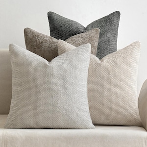 May include: A collection of textured throw pillows in various neutral tones, including light gray, beige, and dark gray. The pillows are arranged on a light-colored sofa, showcasing their woven fabric and square shape. The pillows add a touch of texture and comfort to any living space.