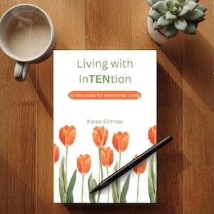 May include: A book titled "Living with InTENtion" with orange tulip illustrations and the text "10 Key Areas for Intentional Living." A black pen rests on the book. A mug of coffee and a succulent plant are also visible.