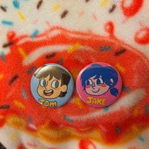 May include: Two round cartoon character buttons. One is light blue with a cartoon boy and the name "TOM" in yellow. The other is pink with a cartoon girl and the name "JAKE" in yellow. The background is a red and white donut.