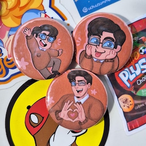 Può includere: Tre bottoni rotondi con illustrazioni di cartoni animati di una persona con gli occhiali e un maglione marrone. I bottoni hanno pose ed espressioni diverse. Altri adesivi e un pacchetto di snack sono sullo sfondo.