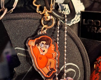 Puede incluir: Un llavero dorado con un dije de personaje de dibujos animados con un atuendo naranja y gafas. El dije está unido a un pompón morado esponjoso. El llavero está sujeto a una mochila negra.