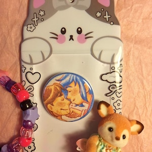 May include: A white cat-shaped phone card holder with gray accents, a round button with a couple kissing, a beaded bracelet, and a small deer figurine wearing a dress. The background is pink.