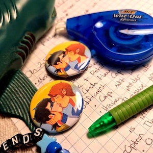 May include: An assortment of items including two round buttons with a cartoon illustration of two people kissing, a blue correction tape dispenser, a green pen, a shark toy, and a beaded bracelet spelling "ENDS".