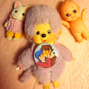 Può includere: Una collezione di giocattoli vintage, tra cui una scimmia di peluche lavanda con un bottone a fumetti, un coniglio bianco con camicia a quadri rosa e pantaloni gialli e una bambola arancione.