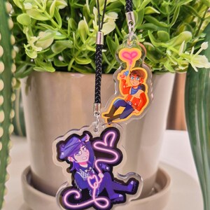 May include: Two acrylic charms with black cords. One charm features a cartoon character in a red and orange outfit holding a ray gun with a pink heart-shaped beam. The other charm depicts a character in a purple outfit with a neon pink design.