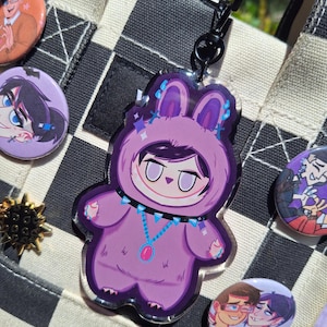 May include: A purple bunny-shaped acrylic charm with a character design, attached to a black metal clasp. The charm features a character in a bunny costume with a blue and pink necklace. Other character pins and a checkered bag are in the background.