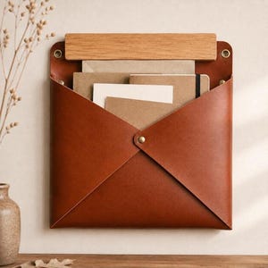 Pode incluir: Um organizador de parede com um bolso de couro castanho em forma de envelope, preso com um botão de pressão central em latão. Uma barra horizontal de madeira clara está fixada no topo com rebites de latão, segurando cadernos e papéis.