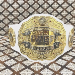 Könnte beinhalten: Ein goldener und weißer Wrestling-Meisterschaftsgürtel mit dem Text "Intercontinental IWGP Champion". Der Gürtel hat aufwendige goldene Details, rote Akzente und einen weißen Lederriemen. Die Seitenteile sind ebenfalls goldfarben.