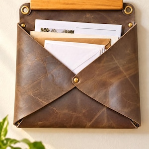 Può includere: Un organizzatore di posta da parete in pelle marrone scuro con una barra superiore rettangolare in legno, occhielli in metallo color ottone e rivetti. La tasca a forma di busta contiene diversi documenti in carta.