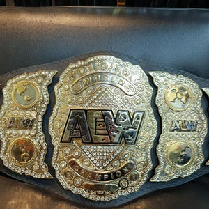 May include: A gold-plated AEW World Championship wrestling belt featuring a large central plate with the text AEW WORLD CHAMPION, surrounded by clear rhinestones. The belt includes two side plates with circular gold medallions and AEW logos.