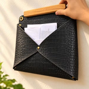 Può includere: Un portacorrispondenza da parete nero con motivo testurizzato effetto pelle di coccodrillo, una maniglia superiore rettangolare in legno e rivetti metallici color ottone. Il supporto a forma di busta contiene documenti cartacei bianchi.