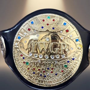 Könnte beinhalten: Ein goldfarbener Meisterschaftsgürtel mit einer großen runden Mittelplatte, die den Text IWGP CHAMPION INTERNATIONAL WRESTLING GRAND PRIX trägt. Die Platte ist mit zahlreichen kleinen, klaren, roten, blauen und grünen Edelsteinen besetzt.