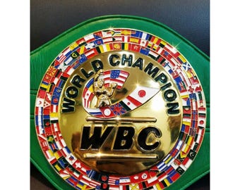 Op de afbeelding: Een gouden WBC World Champion boksriem met een groene leren band. De riem heeft een centraal embleem met een bokserfiguur en vlaggen van verschillende landen langs de rand. De tekst "WORLD CHAMPION" en "WBC" zijn prominent aanwezig.